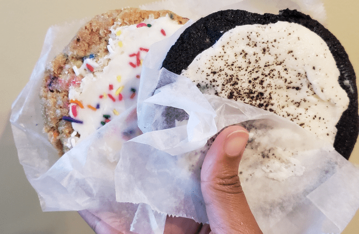 Hand holding a Schmackary's Funfetti Sugar cookie with vanilla frosting and sprinkles and a cookies and cream cookie with vanilla cream cheese frosting and stuffed with vanilla cream cheese frosting from New York City in Hell's Kitchen