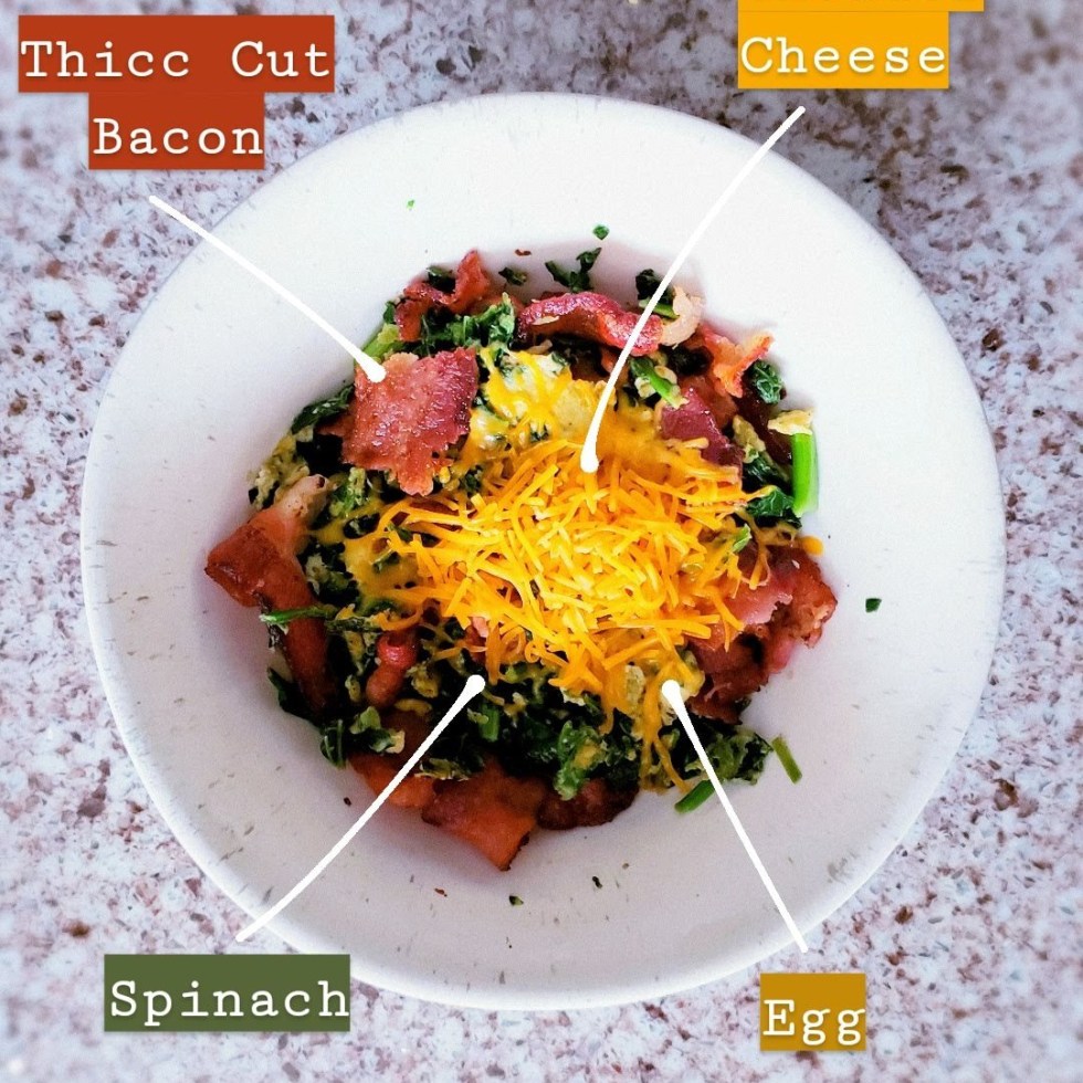 A white bowl and a granite top with Spinach, egg, thick cut bacon and cheddar cheese combined to make a breakfast bowl